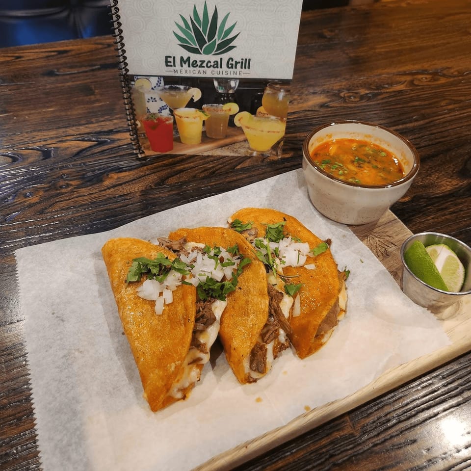 Three El Mezcal Grill birria tacos with a side of consommé and lime wedges on a wooden table.