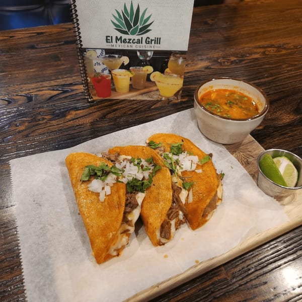 Three El Mezcal Grill birria tacos with a side of consommé and lime wedges on a wooden table.