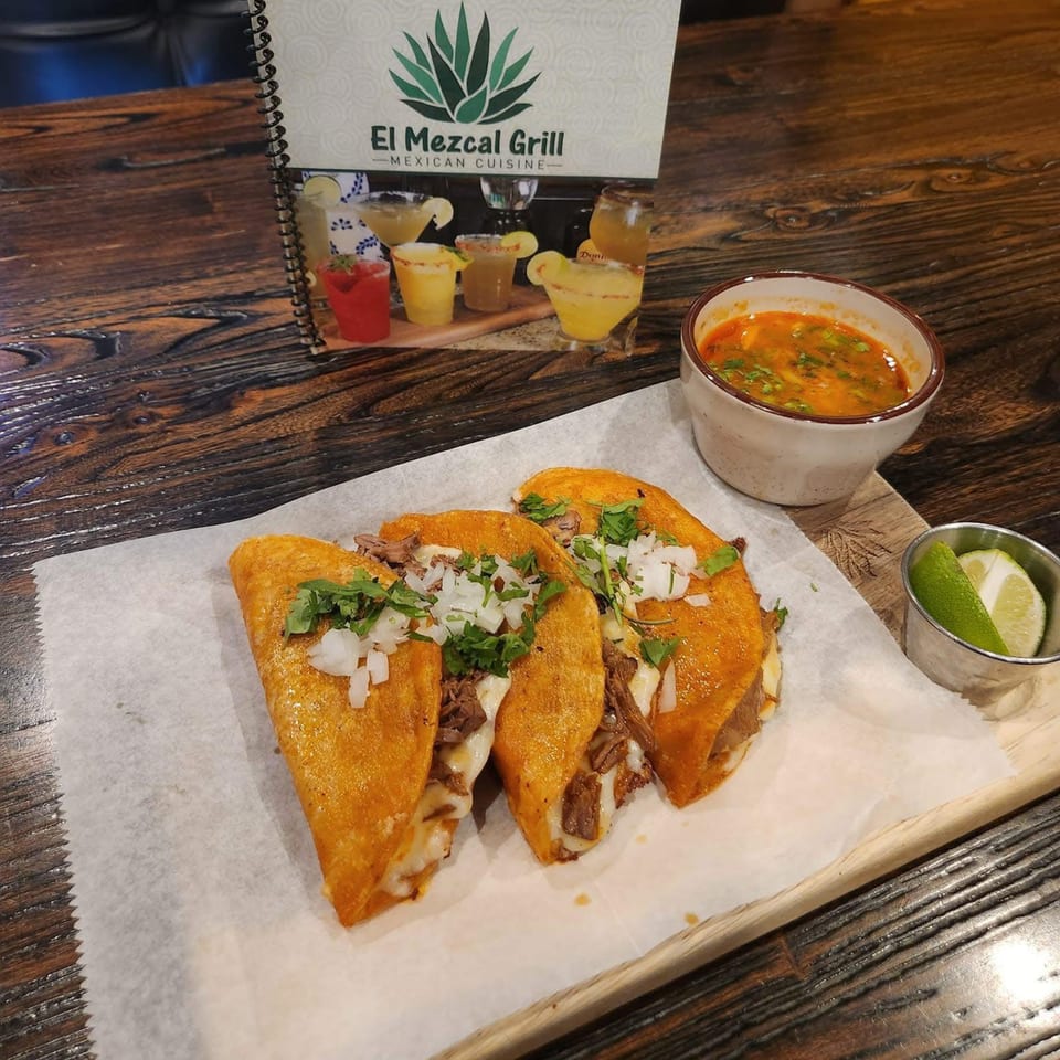 Three El Mezcal Grill birria tacos with consome and lime wedges on a wooden table.