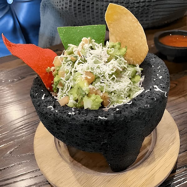 Guacamole in a black stone molcajete with colorful tortilla chips and salsa on a wooden table.