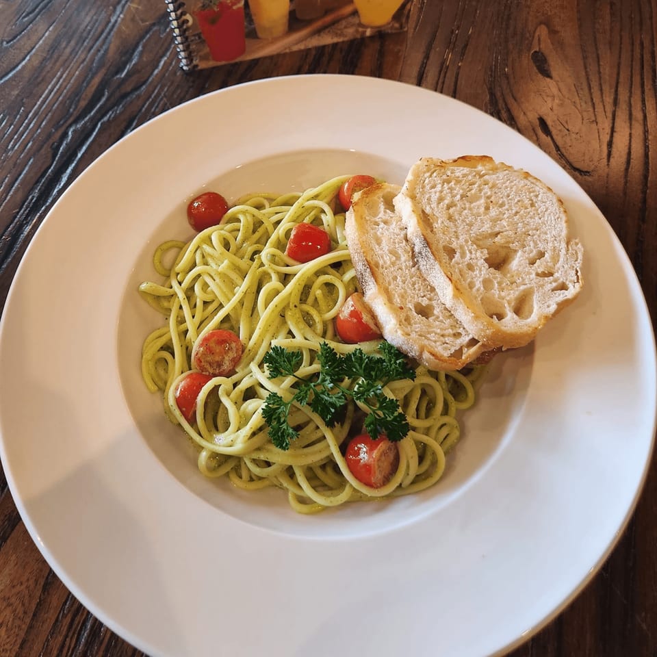Green pesto spaghetti with cherry tomatoes, parsley, and two slices of bread on a white plate.